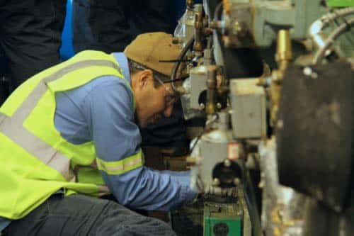 A worker in a high-visibility vest, blue shirt, and brown cap is crouched and focused on inspecting industrial machinery, ensuring reliable turnkey refrigeration solutions in a busy factory setting.