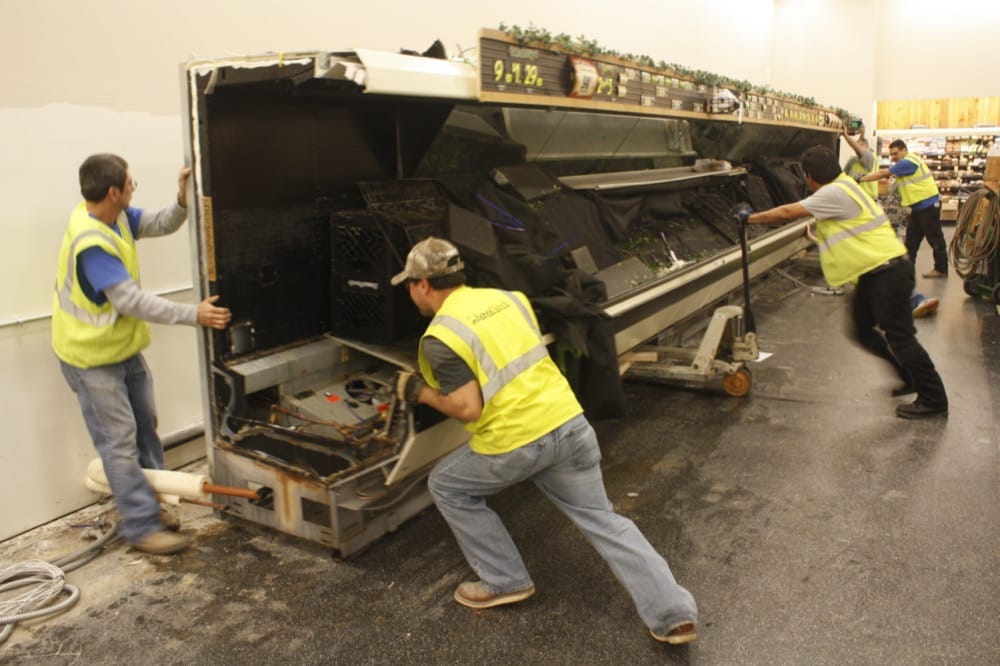 Three workers in yellow safety vests are moving or repairing a large, empty refrigerated display case inside a store, possibly as part of turnkey refrigeration solutions. The case is tilted and the floor around them appears wet.
