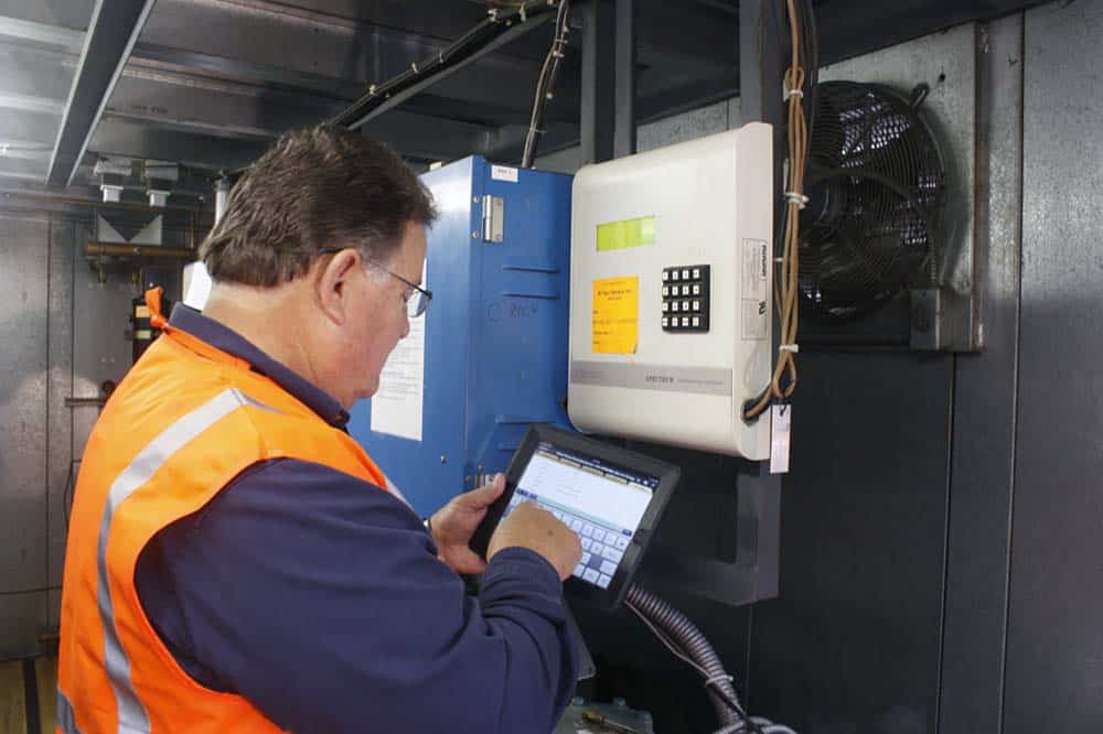 A man wearing a high-visibility orange vest uses a touchscreen device in an industrial setting, standing near electronic control panels that help manage energy-efficiency and modern refrigeration solutions.