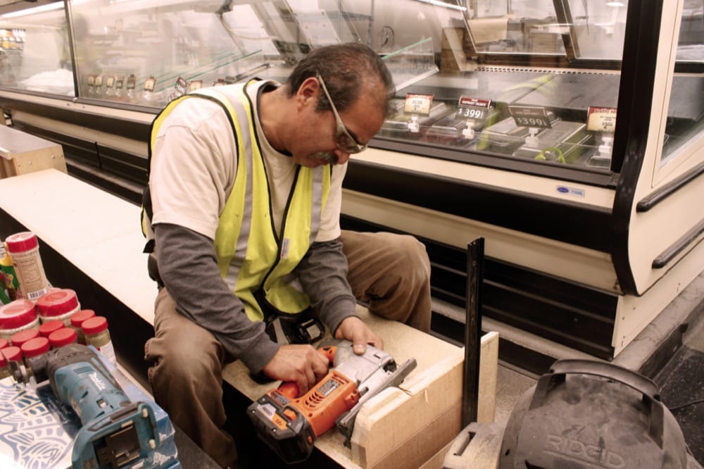 A man wearing a safety vest and glasses uses a power tool to work on a wooden structure inside a supermarket near refrigerated display cases, showcasing turnkey refrigeration solutions. Various tools and materials are scattered around him.