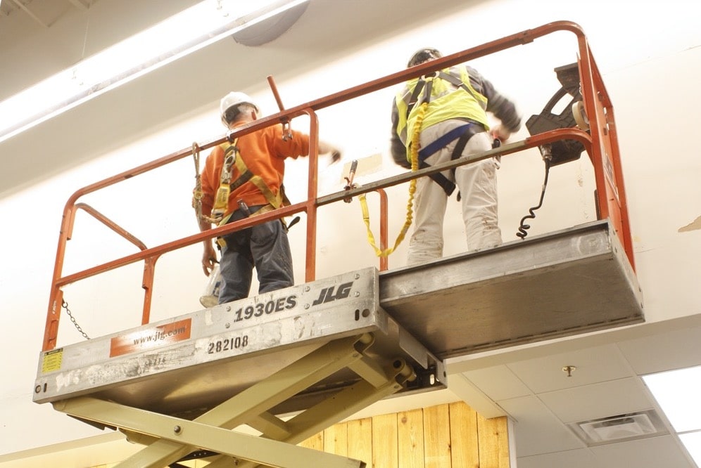 Two construction workers wearing safety harnesses stand on a scissor lift, working near the ceiling of an indoor space equipped for turnkey refrigeration and cold-storage environments, with tools and equipment visible around them.