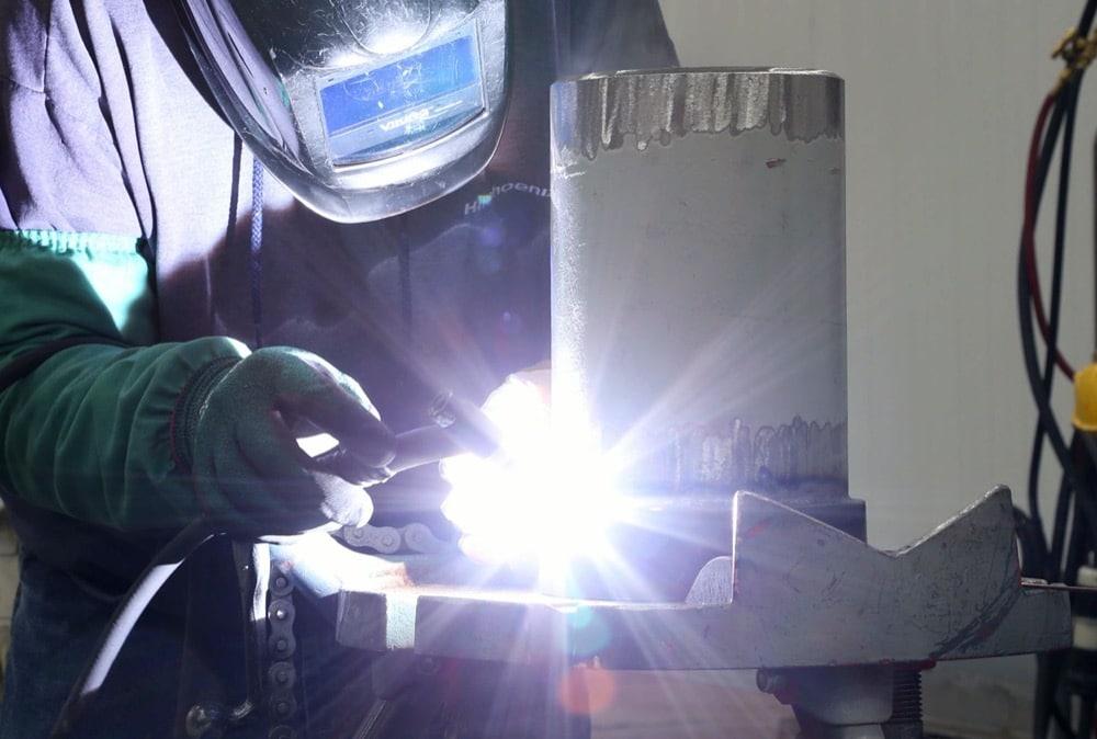 A welder wearing protective gloves and a helmet uses a welding torch on a large metal pipe, working on energy-efficiency solutions for turnkey refrigeration as bright sparks and light illuminate the scene.