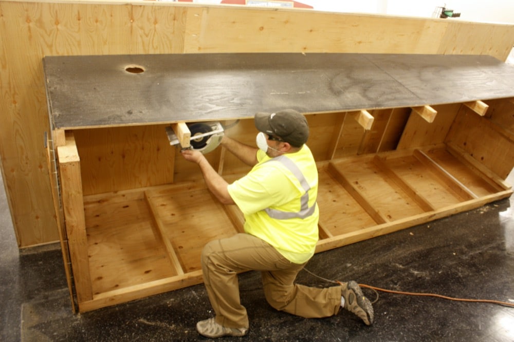 A worker in a yellow safety vest and cap kneels on the floor, using a circular saw to cut wood on a large, partially constructed wooden structure for an energy-efficiency solutions project.