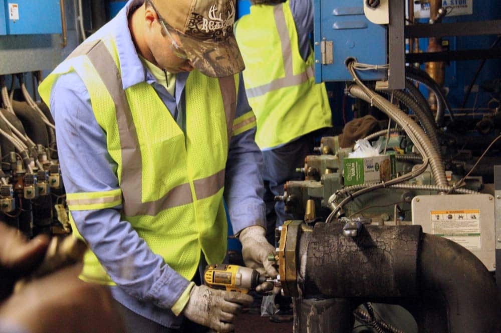 A worker in a high-visibility vest, gloves, and a cap uses a power drill on industrial machinery—possibly installing turnkey refrigeration or energy-efficiency solutions—in a factory or workshop setting. Another worker is visible in the background.