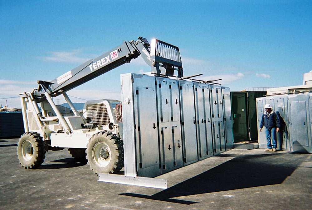 A forklift labeled “Terex” lifts a large metal structure in an outdoor industrial area. A worker in safety gear stands nearby, with equipment related to turnkey refrigeration visible under a clear blue sky.