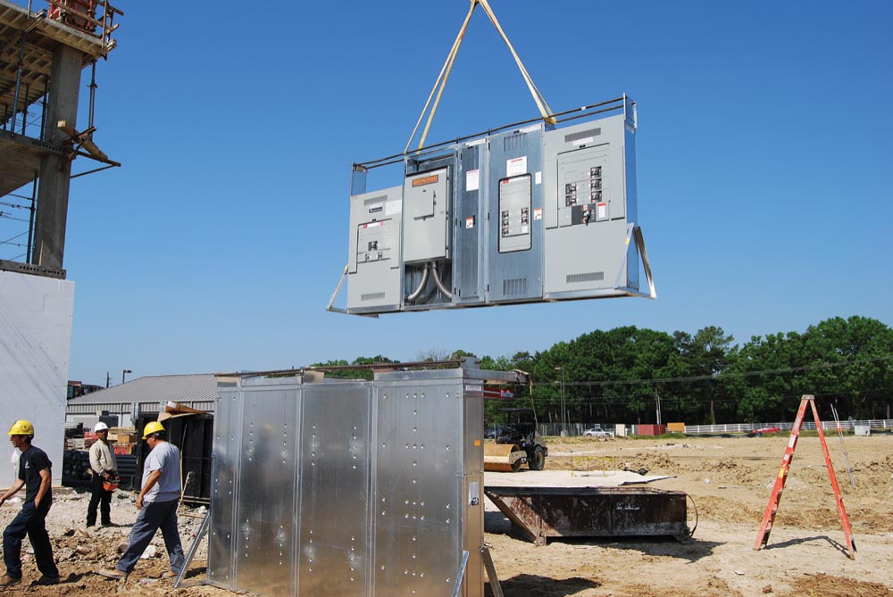 A large electrical control panel for a turnkey refrigeration system is being lifted by a crane at a construction site. Several workers in hard hats observe the operation outdoors, with clear skies and trees in the background.
