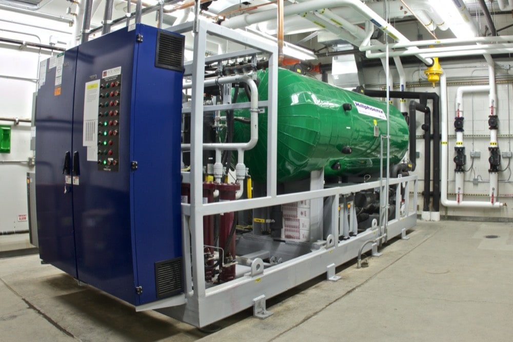 A large industrial refrigeration unit with a green cylindrical tank and blue control panel is installed in a brightly lit mechanical room, providing energy-efficiency solutions for cold-storage with pipes and gauges visible on the walls.