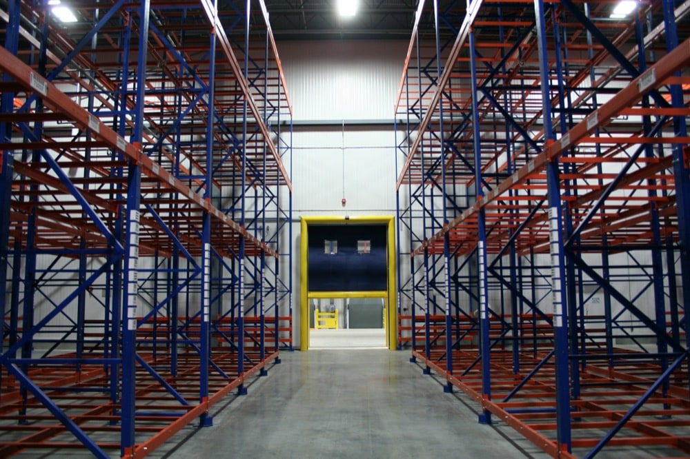 Empty industrial warehouse racking with blue and red metal shelves, forming aisles, and a large industrial door at the end—ideal for implementing turnkey refrigeration solutions, with yellow safety barriers providing added protection.
