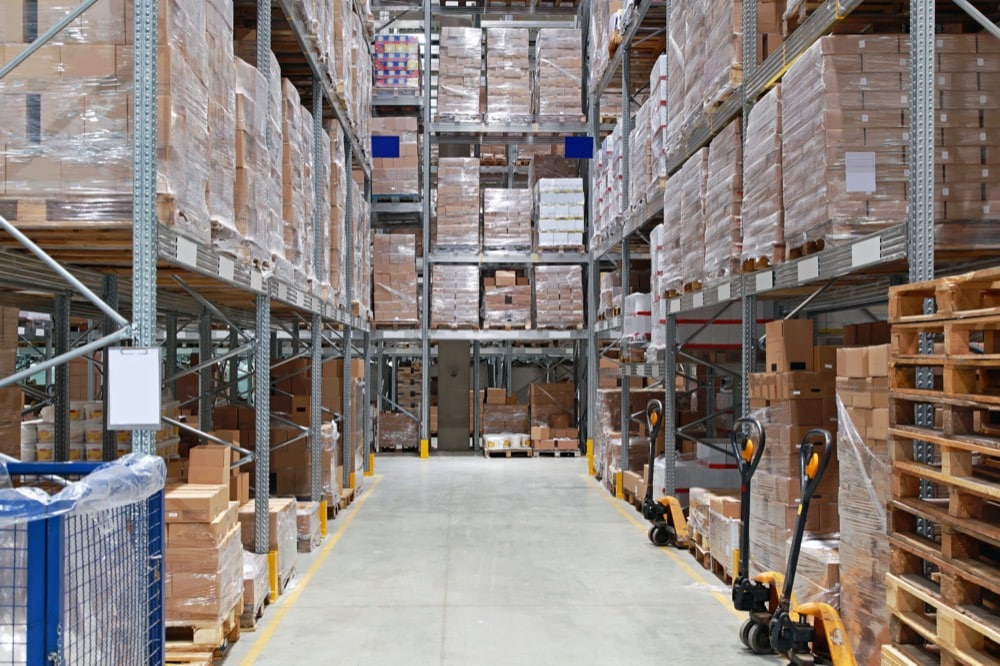 A spacious warehouse with tall shelves stacked with boxes and pallets. Three pallet jacks are visible on the right, and rows of organized goods extend into the background. The well-lit space features energy-efficiency solutions throughout.