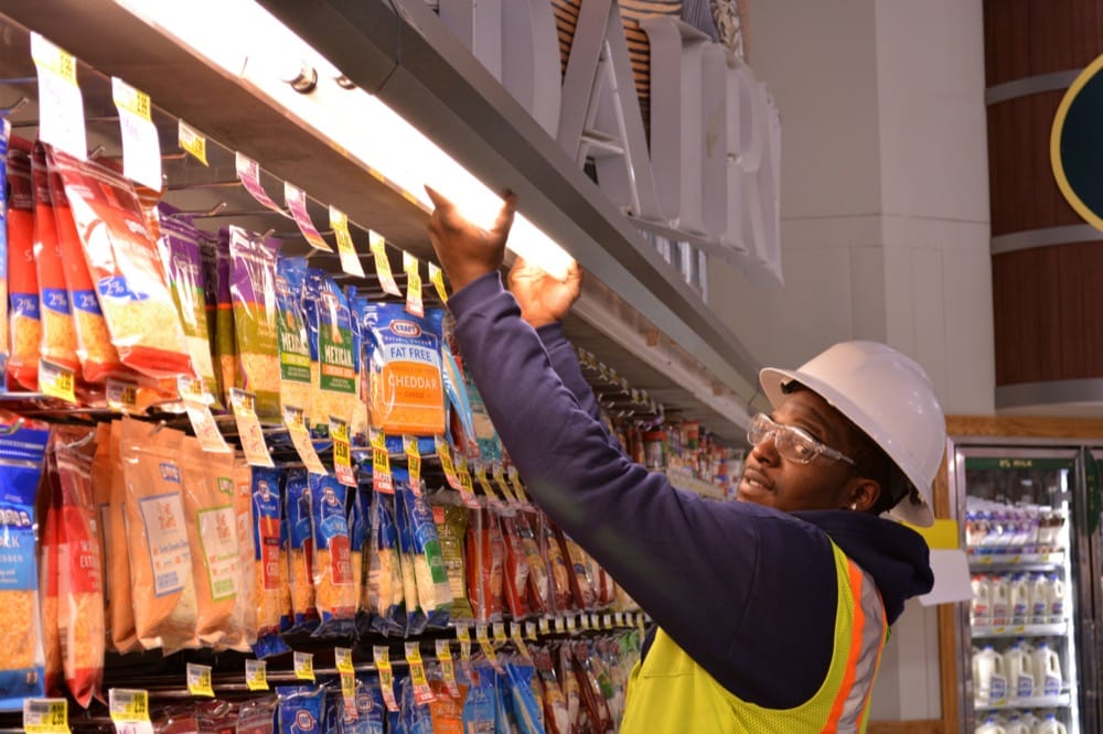 A worker wearing a hard hat, safety glasses, and a reflective vest adjusts a light fixture above shelves stocked with bags of shredded cheese in a grocery store's dairy section, helping implement energy-efficiency solutions.