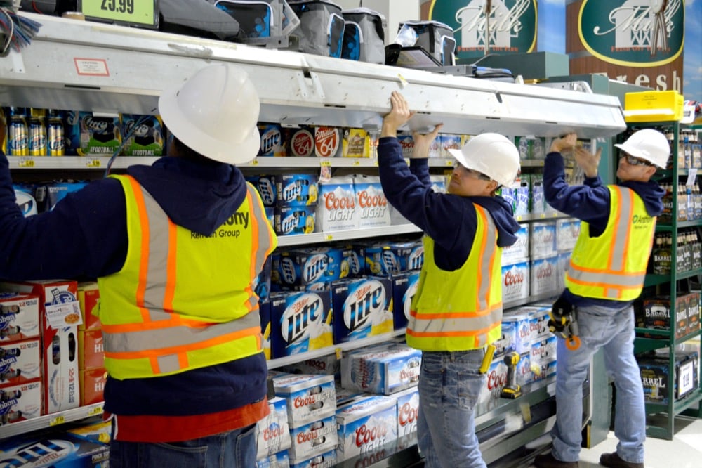 Three workers in safety vests and hard hats are inspecting or repairing a large metal shelf above stocked beer boxes in a store aisle, focused on delivering turnkey refrigeration solutions using specialized tools.