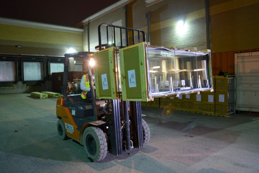 A forklift operator moves a large, crate-wrapped glass window at night outside a warehouse, illuminated by overhead lights, highlighting the facility’s commitment to energy-efficiency solutions.