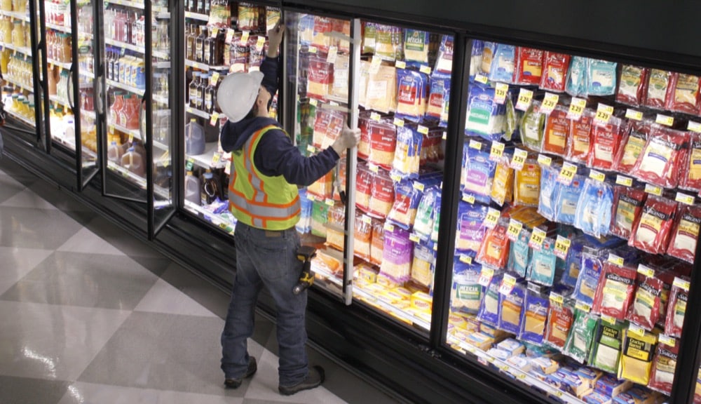 A worker in a hard hat and safety vest stands before refrigerated shelves filled with packaged cheese, reaching into a cooler with a tool—demonstrating turnkey refrigeration solutions in a grocery store setting.