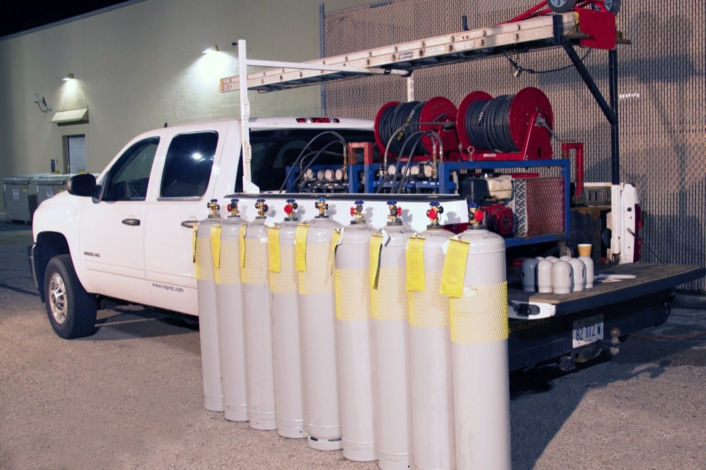 A white utility truck with an open flatbed is parked outside at night, carrying multiple large gas cylinders and equipment, including hoses and gauges—ideal for supporting cold-storage or refrigeration solutions. Some cylinders have yellow tags attached.