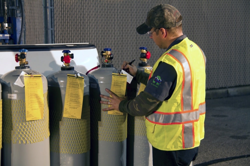 A worker in a yellow safety vest and camouflage cap inspects and writes on tags attached to large gas cylinders, supporting energy-efficiency solutions for refrigeration in cold-storage environments outdoors near a fence.