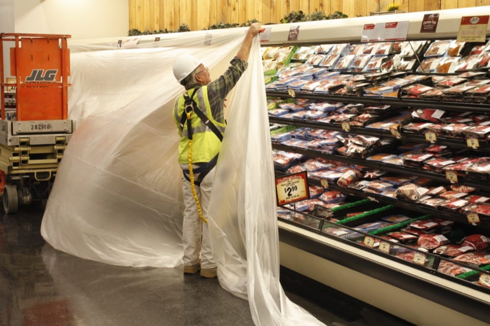A worker in a reflective vest and hard hat hangs clear plastic sheeting in front of a supermarket meat display case, highlighting maintenance in cold-storage environments that demand energy-efficiency solutions.