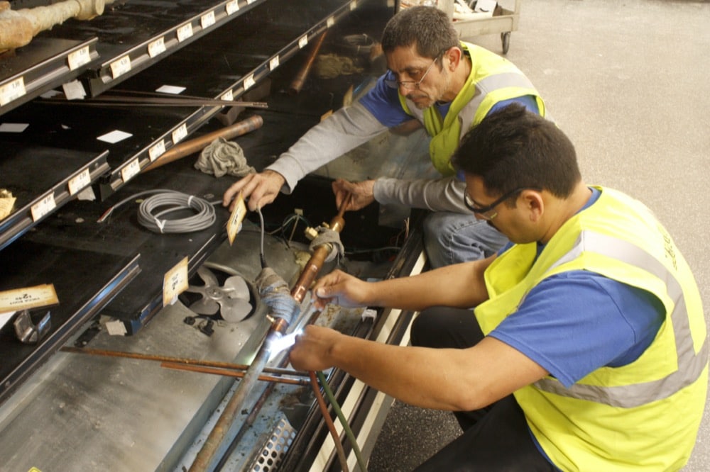 Two workers in safety vests repair metal pipes using a blowtorch and tools, working side by side on a large metal structure—showcasing turnkey refrigeration solutions with a focus on energy-efficiency solutions.