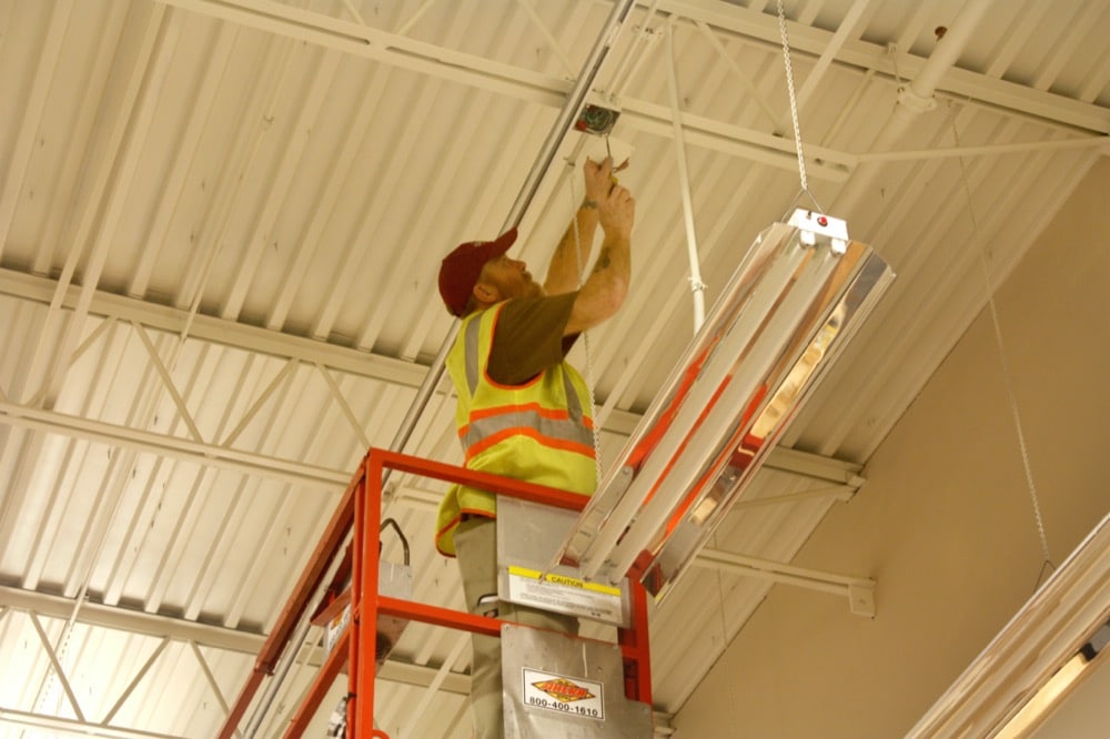 A worker in a safety vest stands on a lift, installing or repairing a light fixture on the ceiling of a commercial building specializing in energy-efficiency solutions.