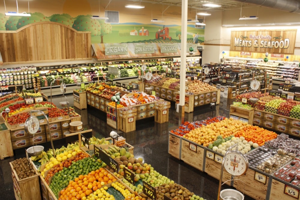 A brightly lit grocery store produce section features colorful fruits and vegetables neatly displayed in wooden bins, maintained fresh with advanced refrigeration solutions, and a sign for meats and seafood in the background.